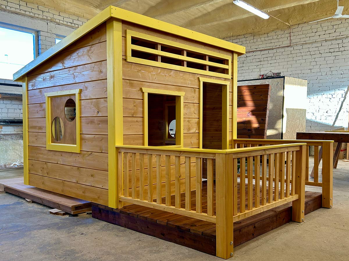 Wooden playhouse with yellow trim, porch railing, and round windows, set up indoors on a workshop floor.