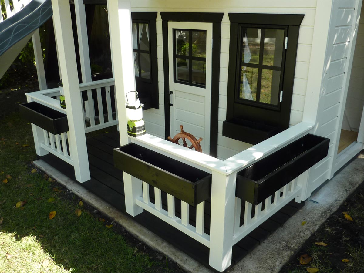 Close-up of a white playhouse porch with a wooden ship wheel, black flower box, and two green camping lanterns.