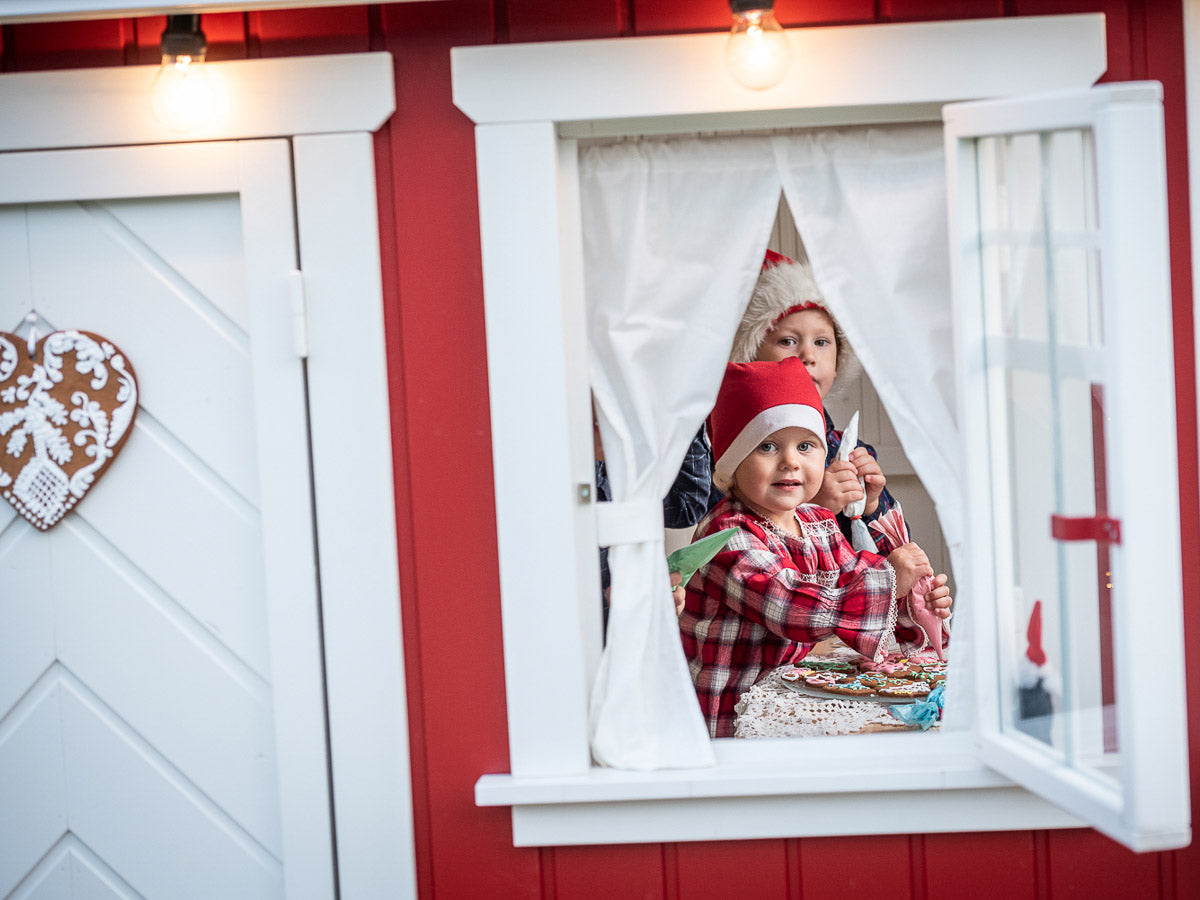 Close-up of the front window of the KidsPlayHouses_EU all-wood red Nordic kids playhouse, with children making gingerbread in the window.