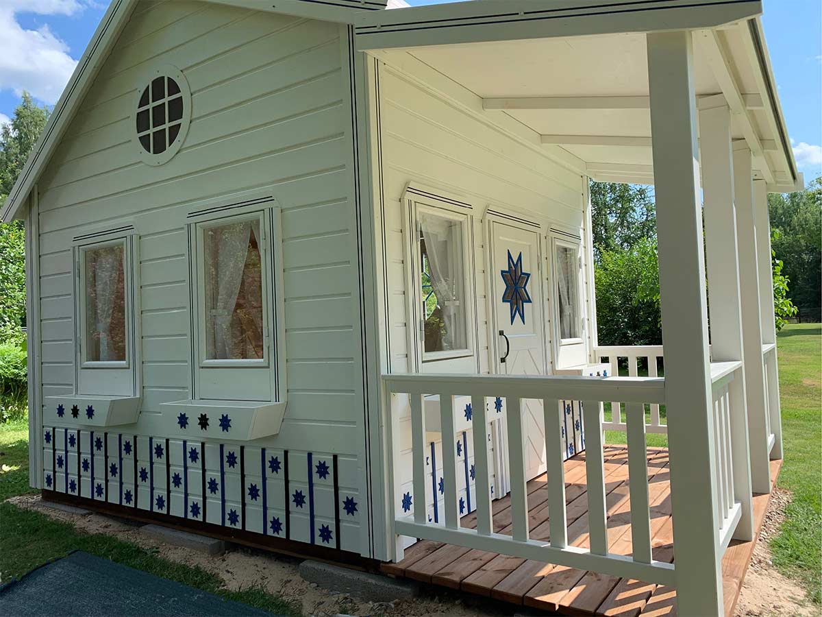 KidsPlayHouses_EU white kids' playhouse Countryside with white-fenced wooden terrace, four terrace posts, roof over terrace and opening windows, close-up view.