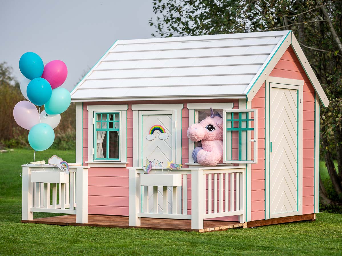 A Unicorn looks out of the open window of the KidsPlayHouses_EU playhouse, and balloons are attached to the terrace railing.