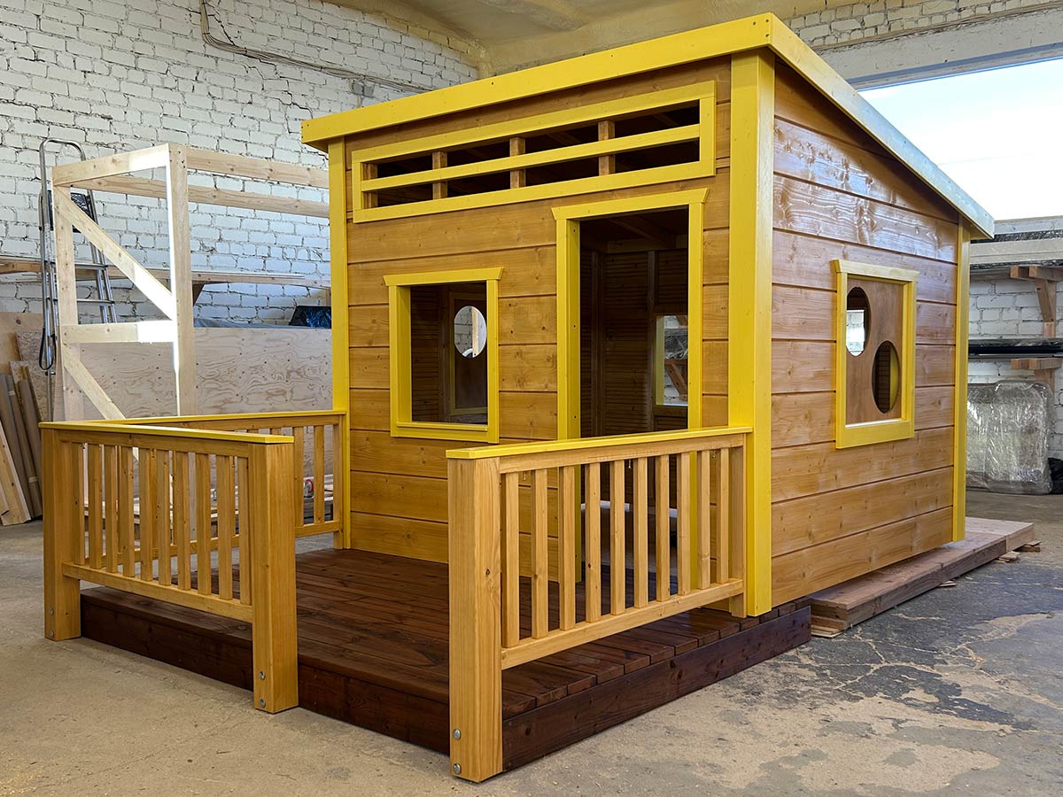Modern wooden playhouse with yellow trim and a slanted roof, displayed indoors on a workshop floor.