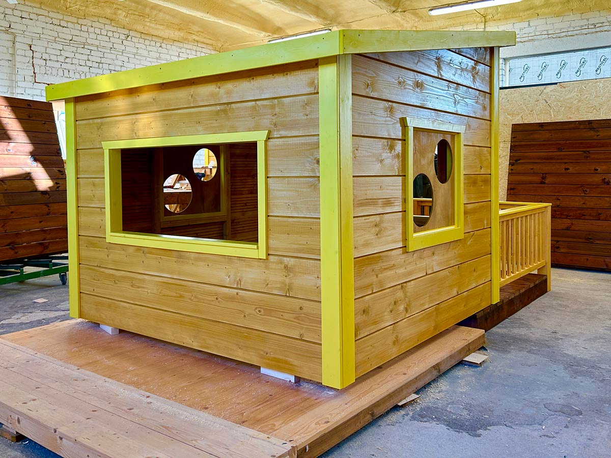 Wooden playhouse with yellow trim and round windows, displayed on a wooden platform inside a workshop.
