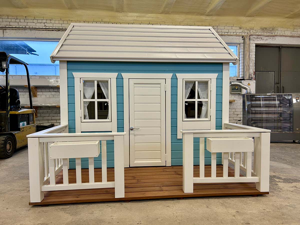 Blue and white wooden playhouse with front porch, flower boxes, curtains and a metal roof, set up indoors.