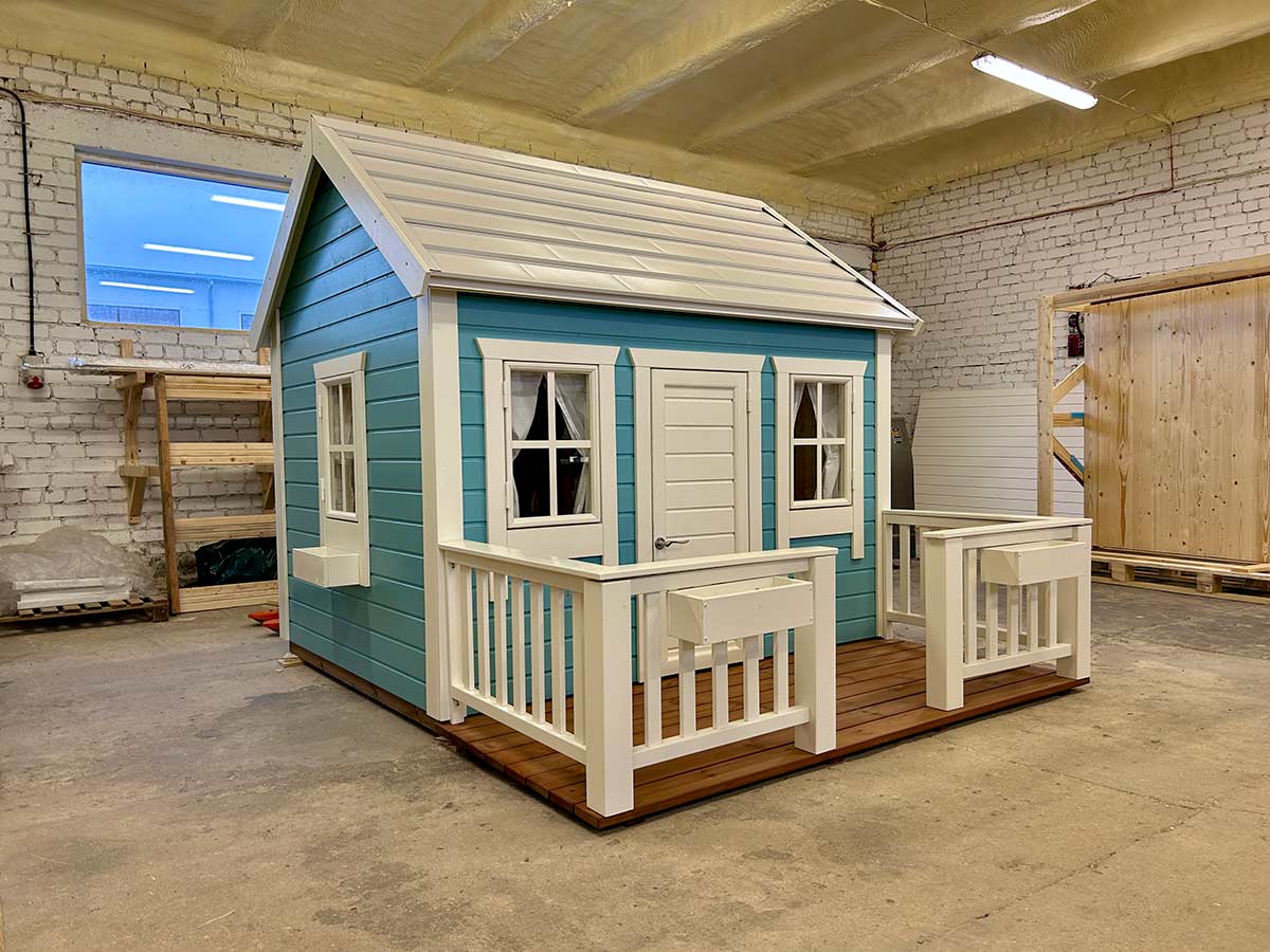 Large pre-painted kids’ playhouse with porch railings, flower boxes, curtains and white metal roof in a warehouse setting.