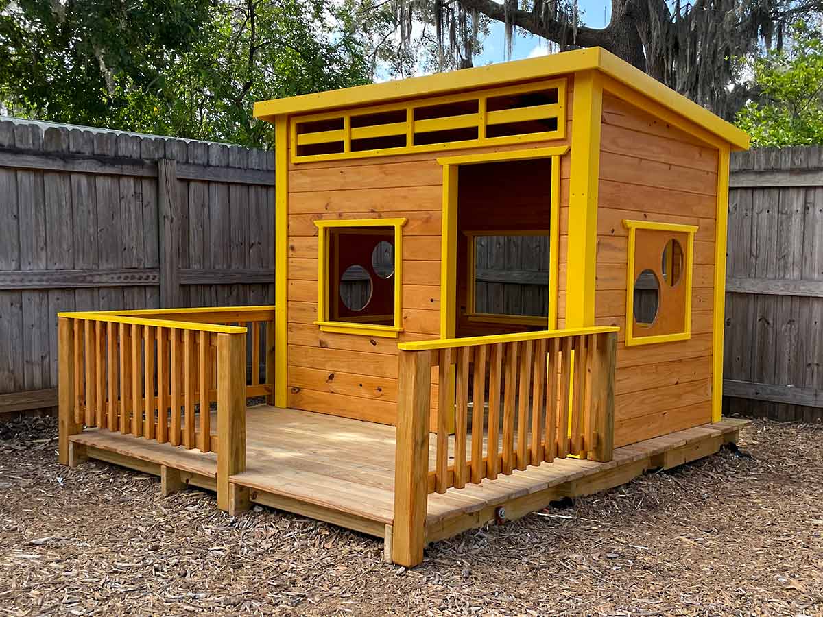 Modern wooden playhouse with a slanted roof, yellow trim, and circular windows, set on mulch in a fenced backyard.