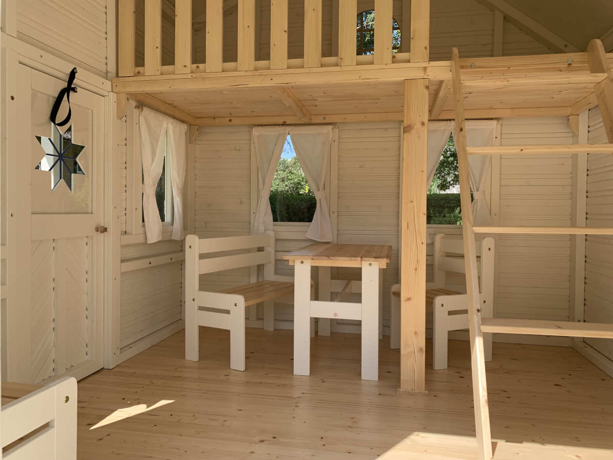 KidsPlayHouses_EU a2 all-wood kids playhouse Countryside with an interior view under the loft, natural wood kids furniture, table, chair and bench. White walls, natural wood floor, loft and ladder to the loft.